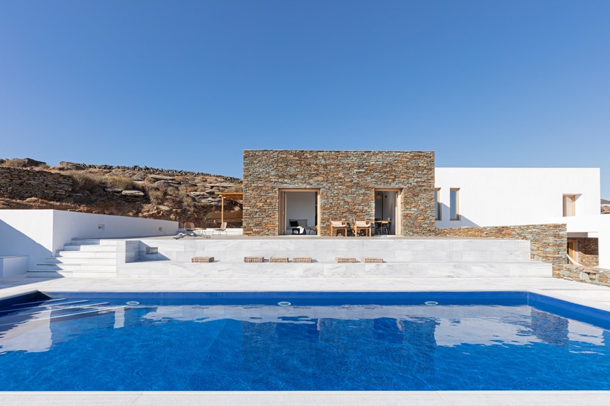 Villa Grace Sifnos — minimalist design with natural stone and white facade, large blue infinity pool, marble deck, and rugged Cycladic hillside landscape