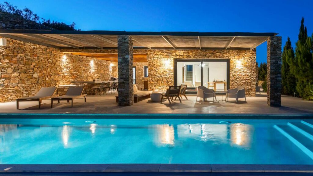 Villa Orchid Sifnos — natural stone facade with covered terrace, lounge seating, and turquoise pool illuminated at evening in Panagia tou Vounou